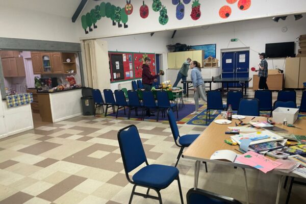 Inside the community centre: a large open step-free space. There is a table with art supplies scattered across it and chairs around it. Part of the room is sectioned off with chairs, and young people are playing ping pong there. The room has decorations on the wall up high of the very hungry caterpillar. There is a kitchen visible to one side.
