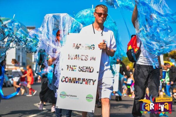 Young people marching down the road with blue jellyfish costumes, one young person holds a large sign reading "making waves in the SEND community"