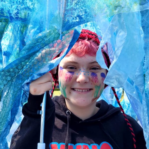 IMG-20230527-WA0005 Young person with jellyfish painted on their face, holding up an umbrella they made into a jellyfish for Eastbourne Carnival. They are surrounded by the jellyfish tendrils in many shades of blue.