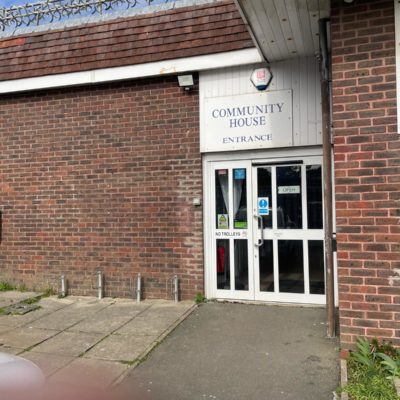 Brick building, with a sign reading "Community House" above the entrance. It is a large door with signs reading "Keep back" and "automatic door". The entrance is flat and wide.