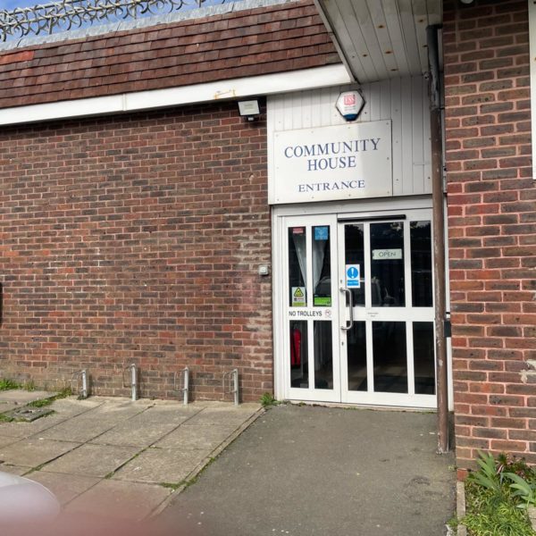 Brick building, with a sign reading "Community House" above the entrance. It is a large door with signs reading "Keep back" and "automatic door". The entrance is flat and wide.
