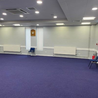 Large room with blank walls and purple carpet. There is a plastic chair by the wall, and radiators.