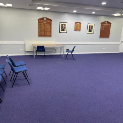 Other side of the same room. There are more chairs and a table. On the walls are two pictures and some wooden panels with engraved names.