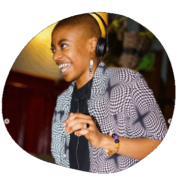 Serena DJing. She is a young Black woman with a shaved head, wearing a shirt with a monochrome psychedelic pattern, and has headphones on. She is looking at someone off camera and laughing.