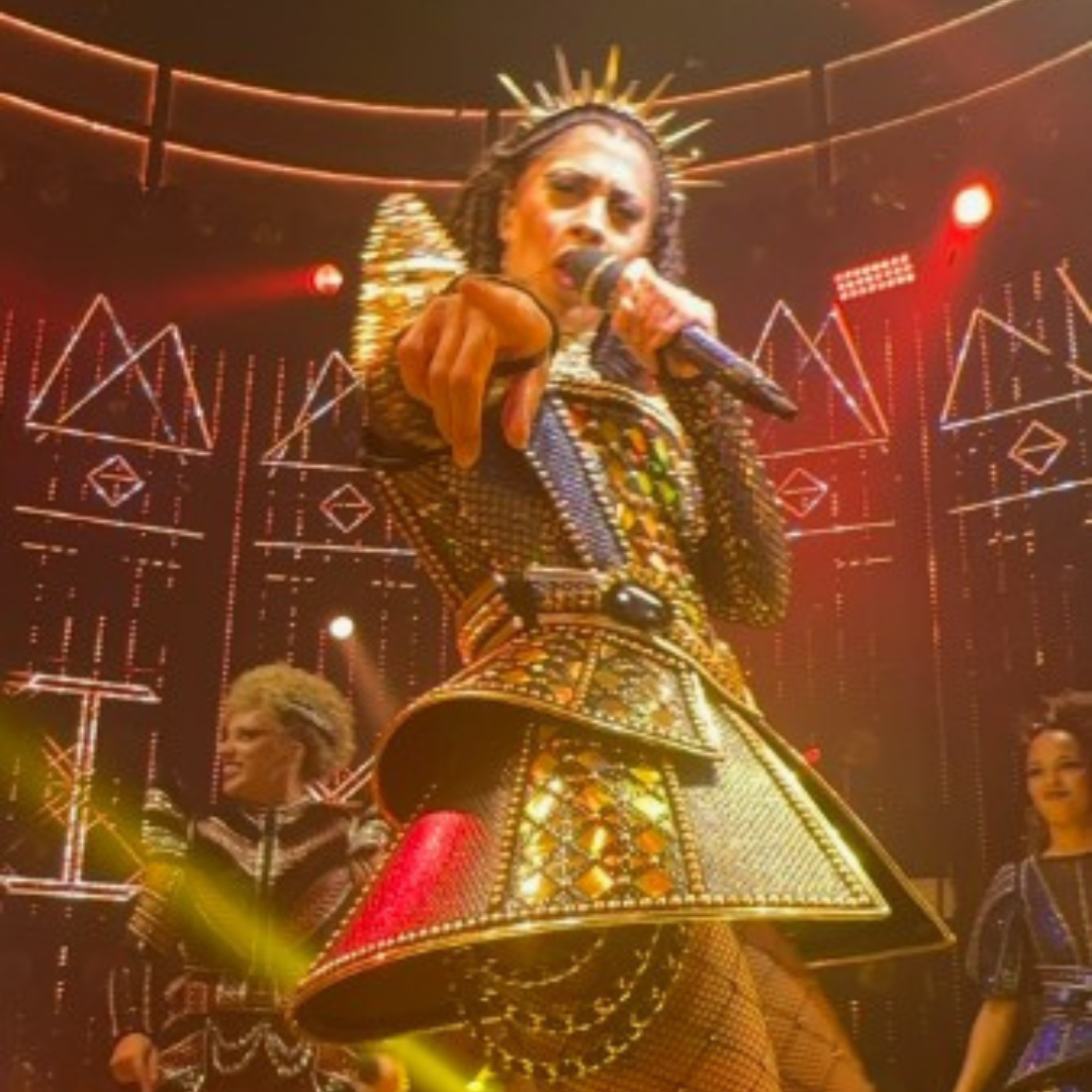 3 Actress holding a microphone and pointing directly at the camera. She has a spiked headdress and a corseted short dress that's a modern take on Tudor dress.