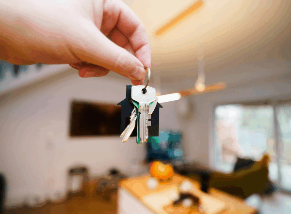 A hand holding a set of keys in front of a blurred background of a living room.