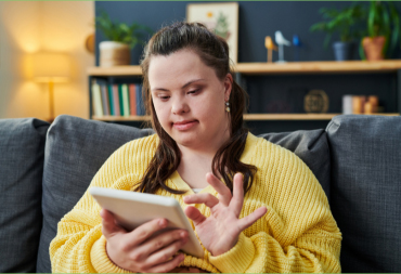 young woman with down syndrome in a yellow jumper sits scrolling on a tablet