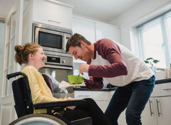 Young woman in a meal chair smiling while her dad helps her with feeding in their white kitchen.