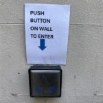 Large button on outside wall with sign reading: "Push button on wall to enter"