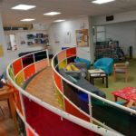 Brighton & Hove Speak Out space. It is on two levels and has a large gentle sloping ramp between the two levels, with brightly coloured transparent panels on the railings around it. There are different kinds of seats, and small tables. There is a pinboard on the wall with notices.
