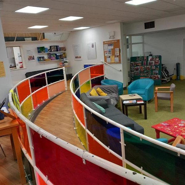 4e304f7d-87b4-488f-a04d-9b75e1f63dab Brighton & Hove Speak Out space. It is on two levels and has a large gentle sloping ramp between the two levels, with brightly coloured transparent panels on the railings around it. There are different kinds of seats, and small tables. There is a pinboard on the wall with notices.