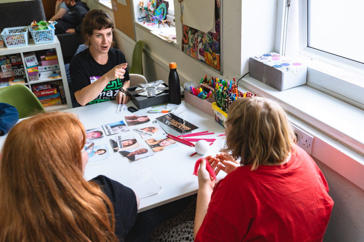Brighton Lead, Sally B, sat at a table with young people, that has photos of Amazing Futures staff and their names. There are lots of art supplies next to them.