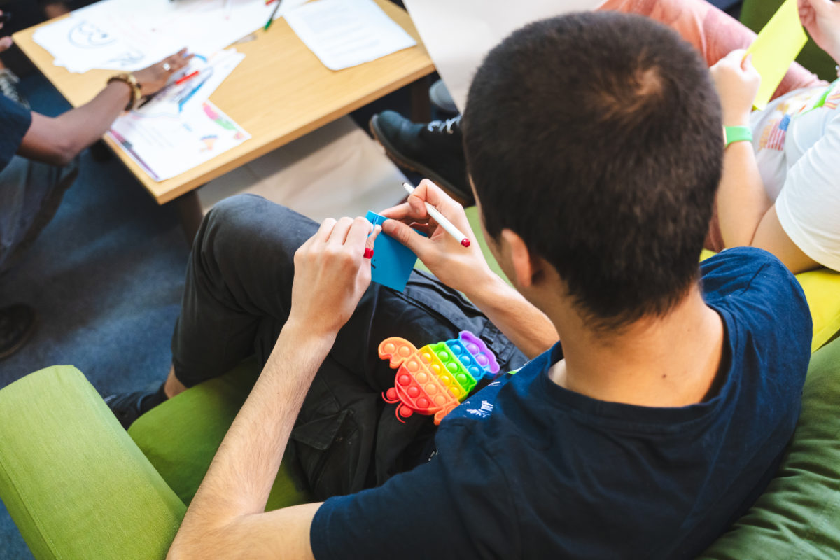 Looking down over a young person's shoulder, they are writing on a pad of post-it notes, and there is a rainbow fidget toy on their lap.