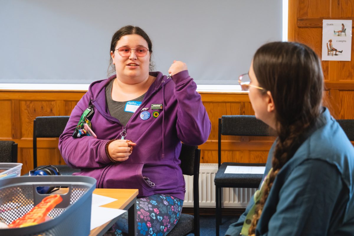 Rose from the Comms Team speaks with a young person, who is wearing a purple hoodie with multiple badges, including a sunflower badge.