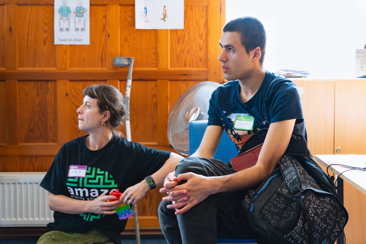 Brighton Lead, Sally B, sat on the floor leaning on a chair next to a young person. They are both watching someone out of view.