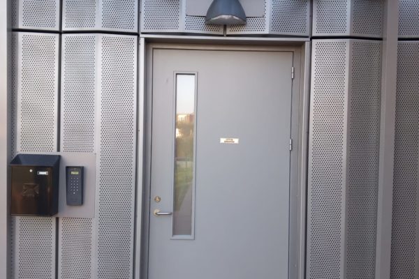 Entrance to the building. The door is wide, with a low handle. There is a buzzer to the left of the door, next to a box for post. The wall is made of metal with a grid of holes. There is a light above the door, and a small roof providing shelter.