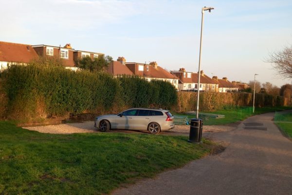 Small parking area with space for a couple of cars, next to a wide path with lampposts. There is a large hedge with houses visible behind.