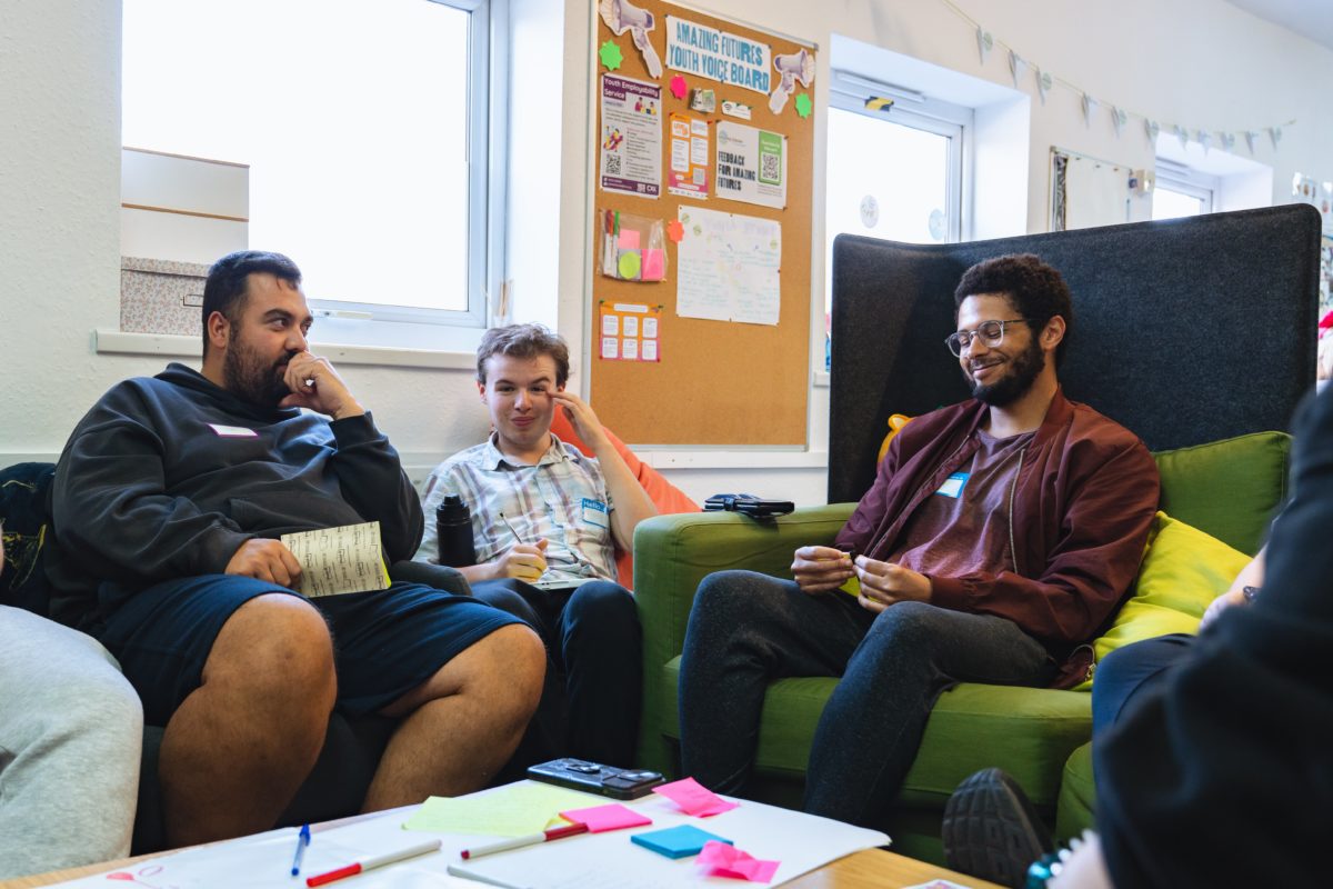 Three young people sat on comfy chairs around a table with post-it notes and pens and papers.