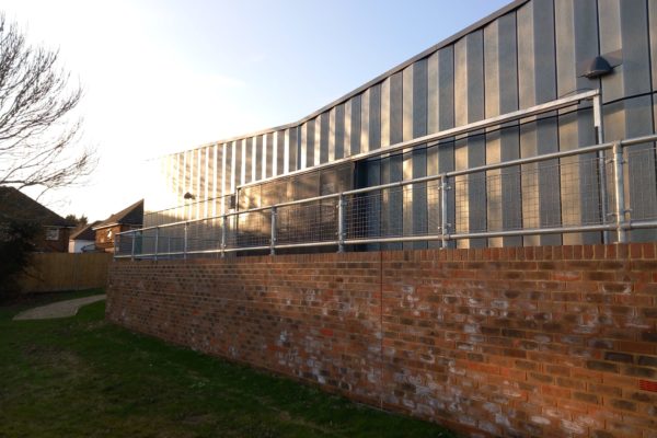 Outside the building, showing the start of the path to the side door. There is a brick wall and the industrial looking building behind. There is grass on the near side of the wall, and at the end of the wall a gravel path begins.