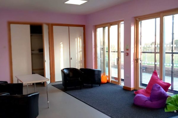 Main space. It's an open space with large glass sliding doors with paving and grass visible beyond. There are colourful beanbags on the floor, a table, and several padded comfy rounded chairs like small arm chairs.