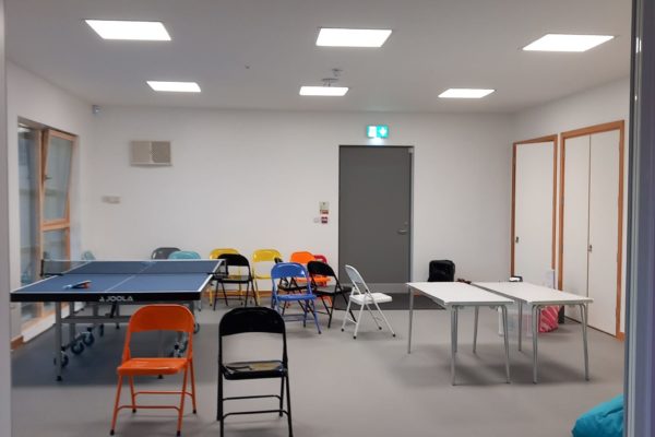 The room is divided into areas. This is the activities space with the ping pong table and folding chairs. There are large windows, and lights in the ceiling.