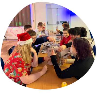 Young people sat around a long table doing a creative activity with newspapers. One is wearing a Santa hat.