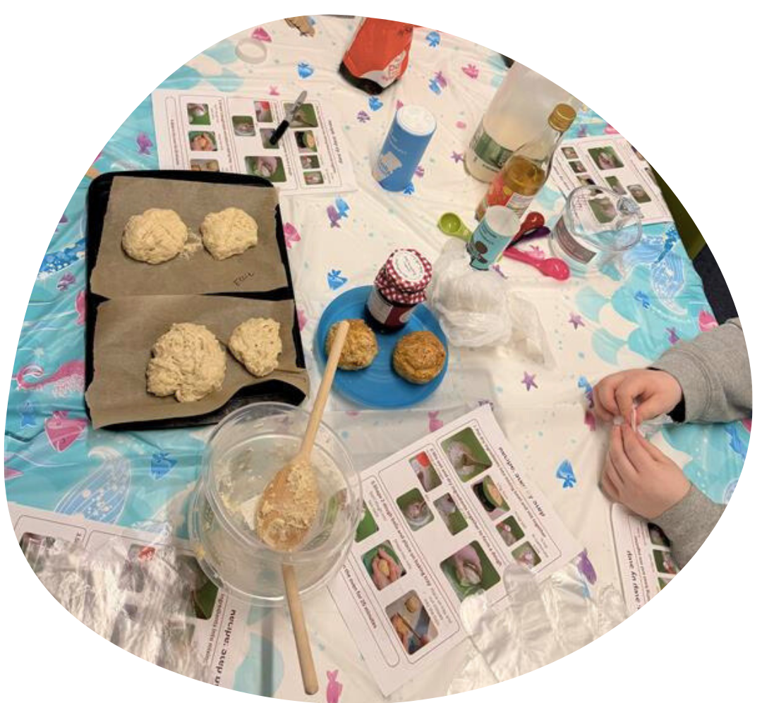 Table with soda bread dough on baking sheets, mixing bowls and wooden spoons, and instruction sheets with lots of photos. A young person is sat at the table, their hands are visible.