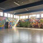 A large sports hall with heavily graffitied walls. There is a basketball hoop and a football net set up at one end.