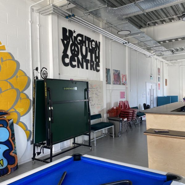 Room with sign on the wall reading "Brighton youth centre" in funky lettering. There is a pool table and a ping pong table visible in the room, and stacks of plastic chairs.