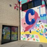 Outside of the building. It has been painted a brightly coloured abstract design, with a large blue letter C. There is a wide glass door and window.