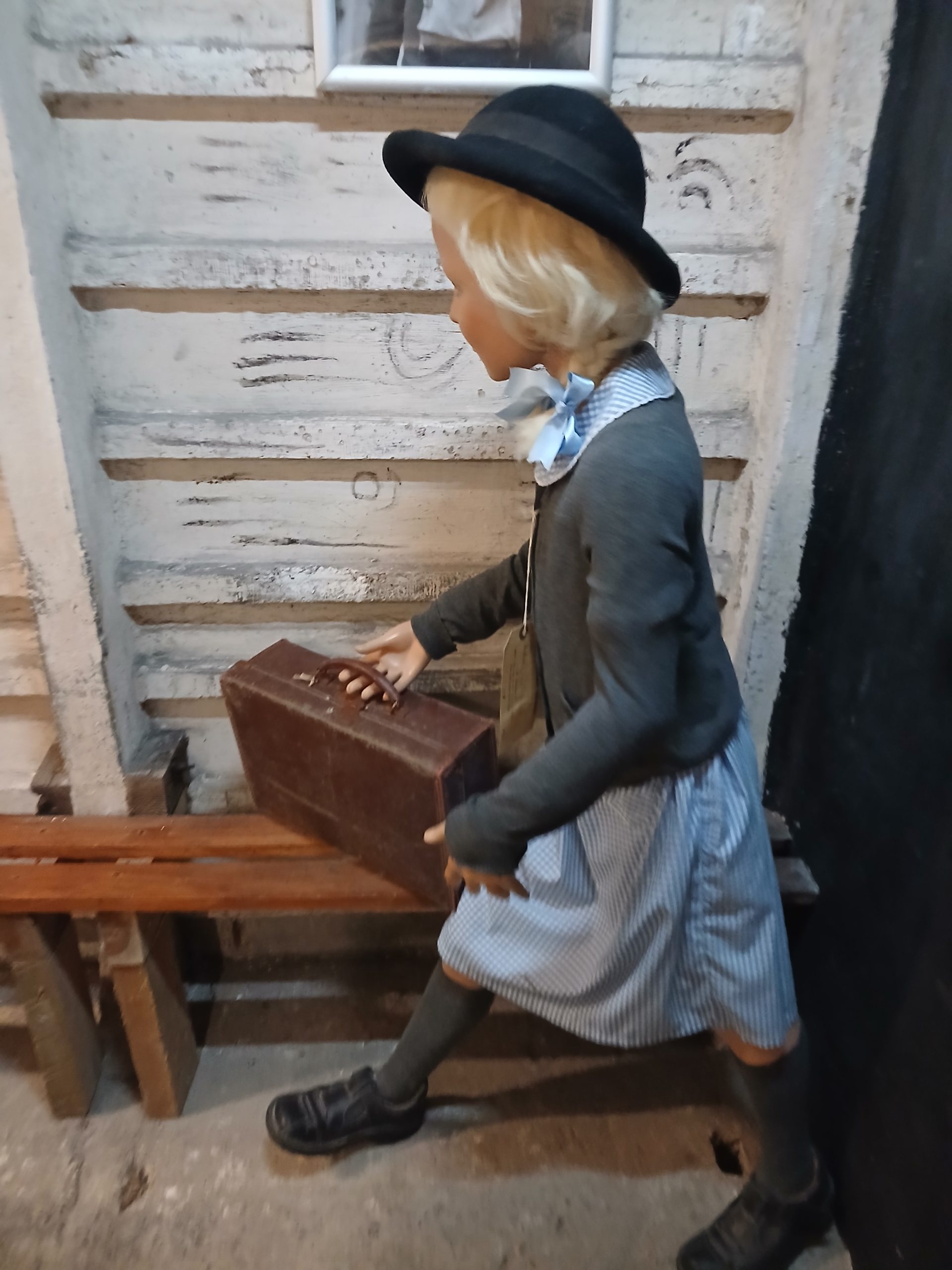 A life size model of a child in 1940s school uniform, holding a small suitcase