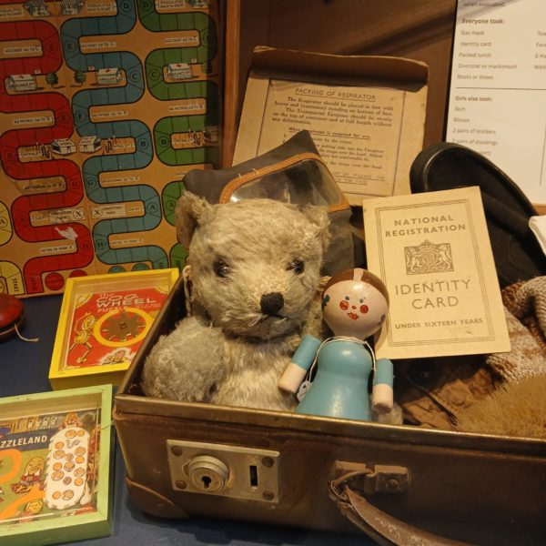 Display showing an evacuee's suitcase, containing a teddy bear and wooden doll, along with an identity card and respirator. There are other vintage children's toys like board games and card games on the table too.