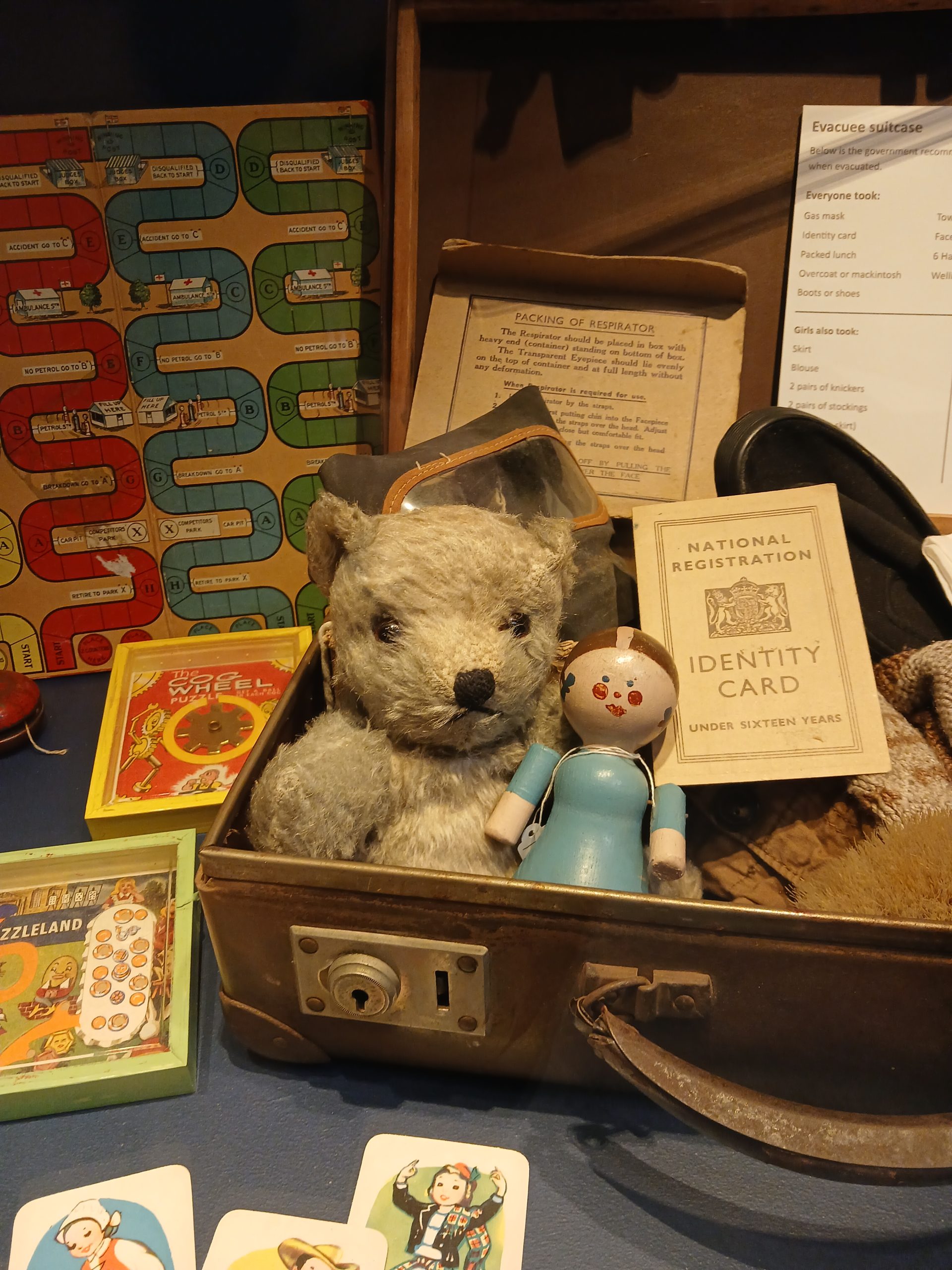 Display showing an evacuee's suitcase, containing a teddy bear and wooden doll, along with an identity card and respirator. There are other vintage children's toys like board games and card games on the table too.