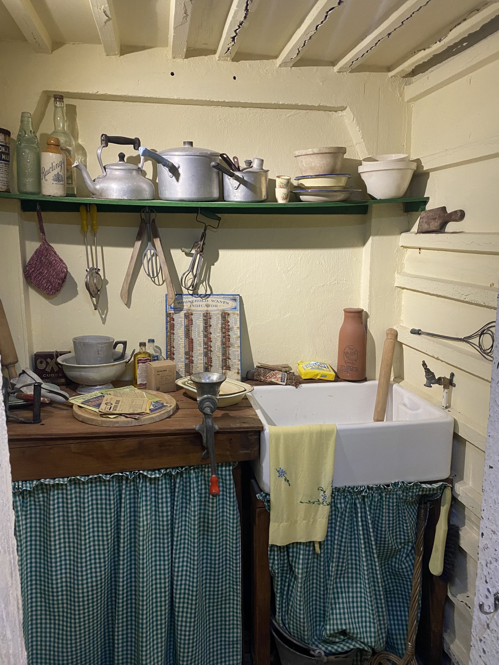 DIsplay showing what a kitchen looked like, with vintage cooking implements on a shelf and table next to a sink.