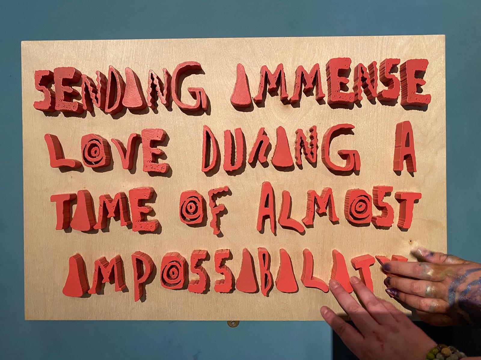 Artwork reading: "Sending immense love during a time of almost impossibility." The words appear cut out of paper, and someone is touching the artwork, feeling the letters.