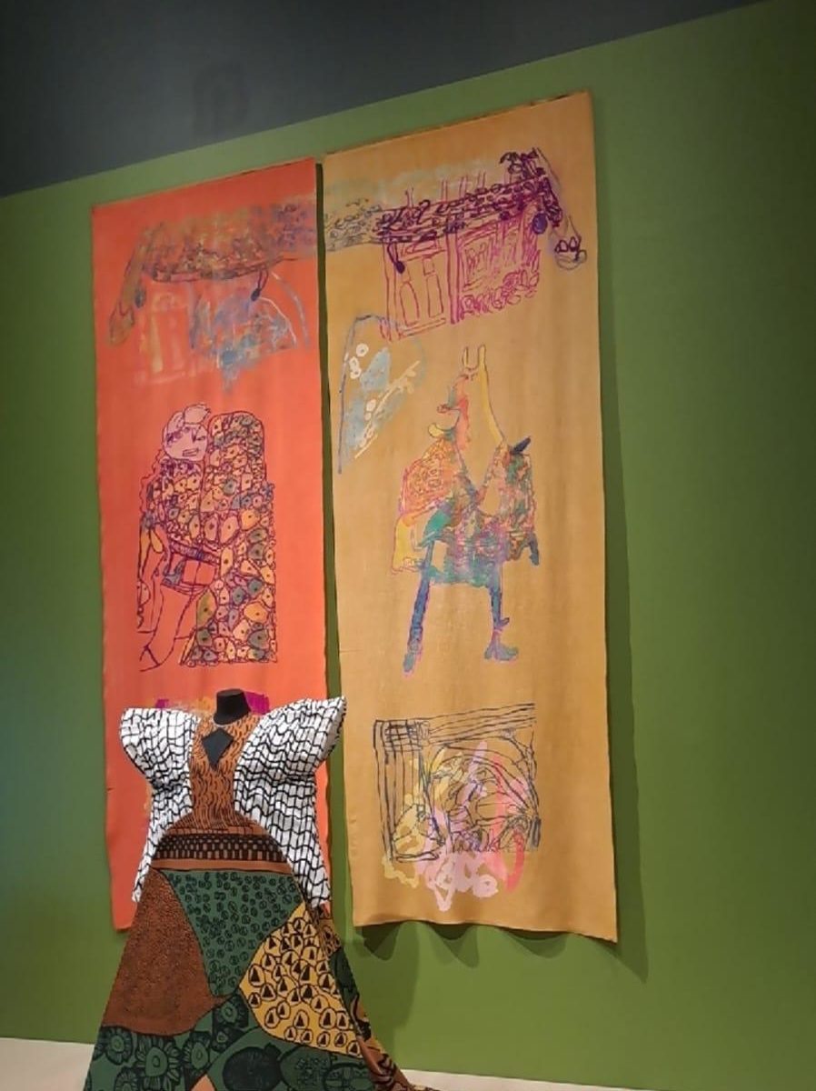 Previously pictured gown, with two banners visible on the wall behind, featuring abstract art in the same style as the art on the dress.