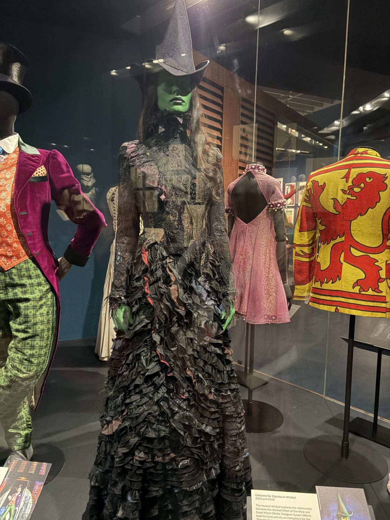 Costumes on display, including Elphaba's costume from Wicked, a long black dress and pointed witches hat, and the model has green skin.