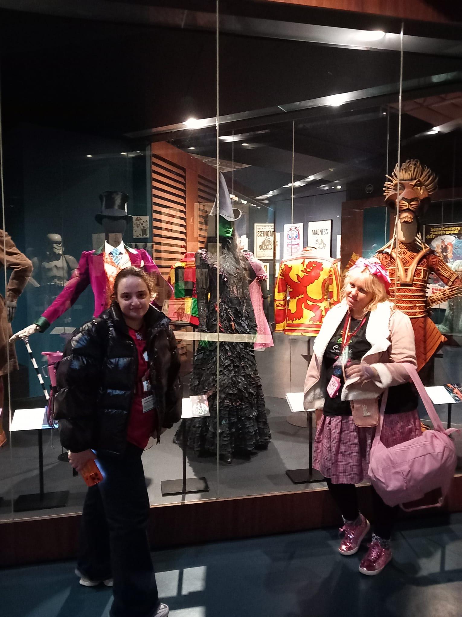 Young people posing for photo in front of a case showing costumes from musicals, including Wicked.