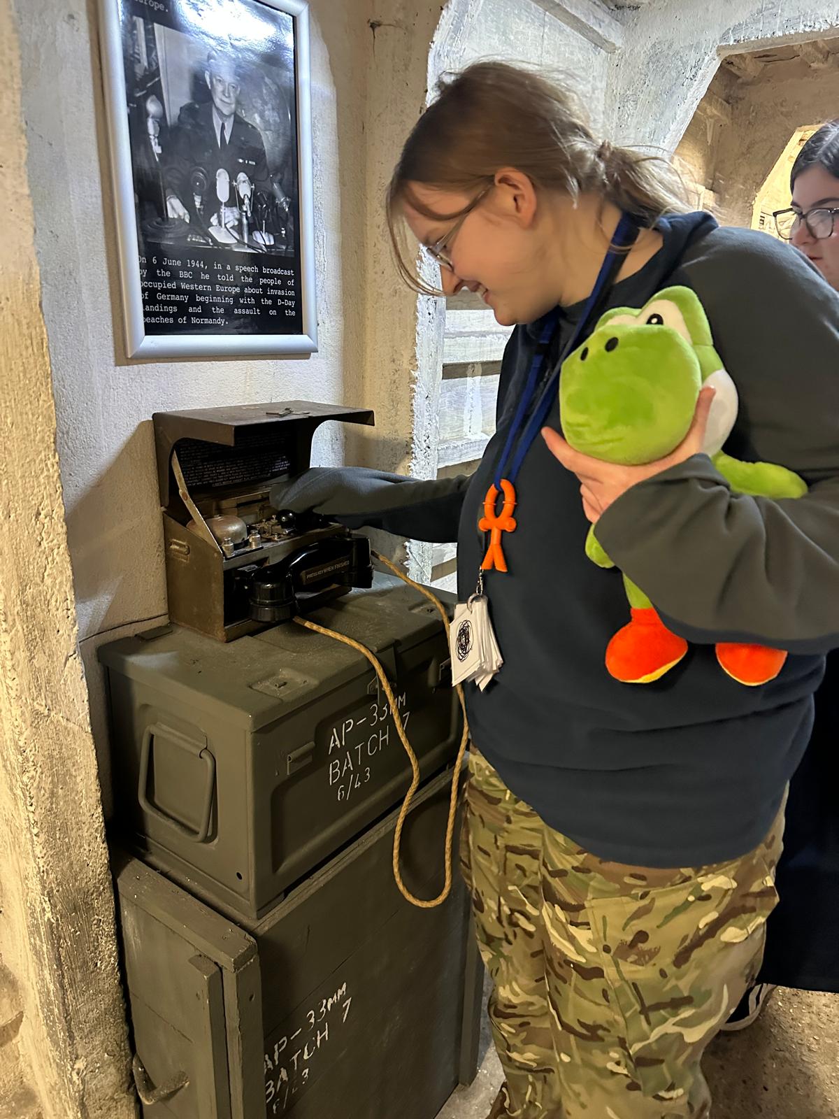 Young person holding a green cuddly toy touching an old military radio, sat on top of military trunks and boxes.