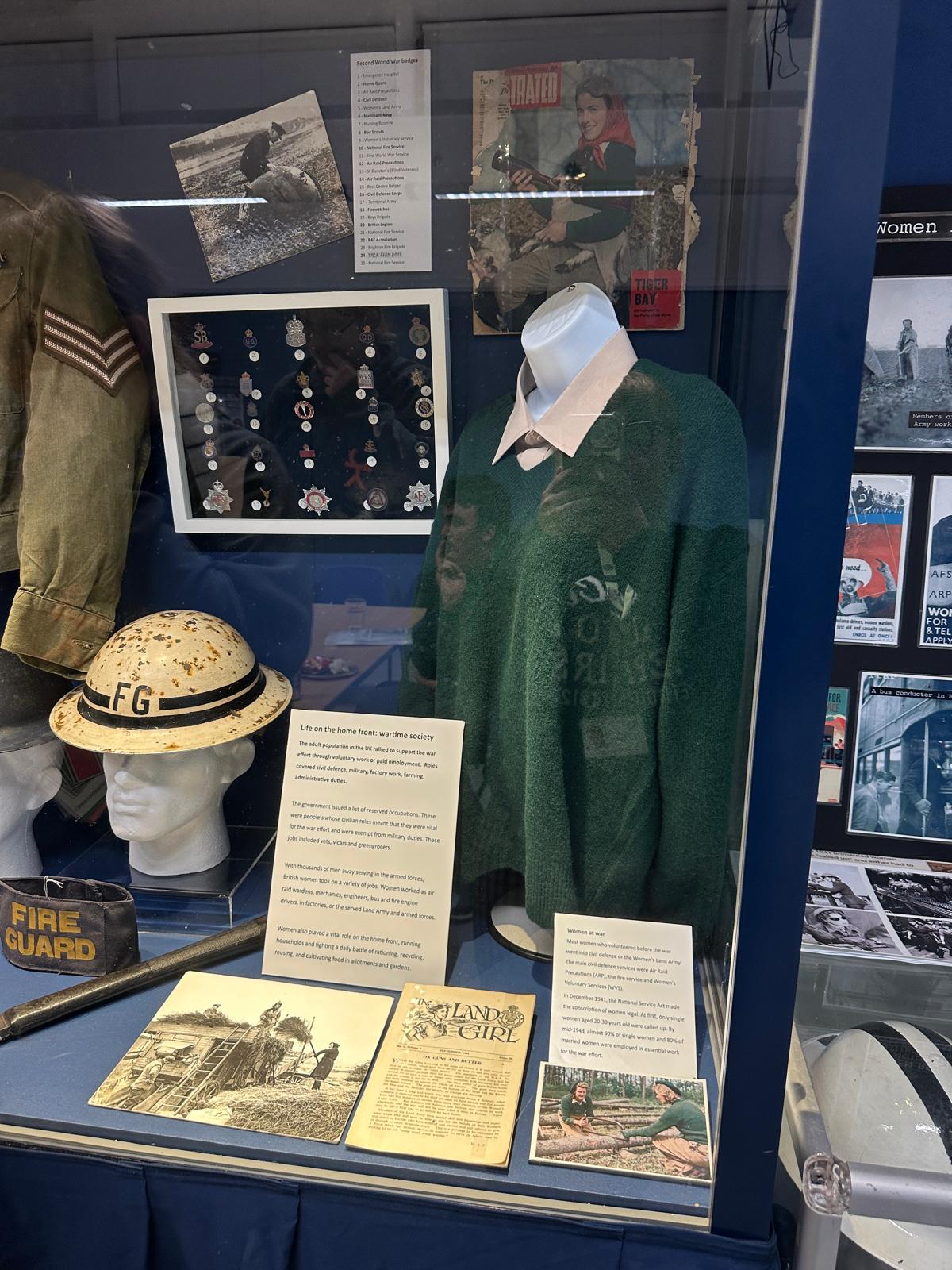 Display showing clothes, including a green jumper, helmet, and military jacket, along with medals and photos.