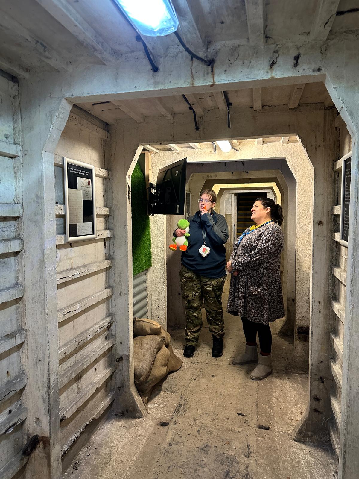 Young person and a staff member watching something on a TV screen, inside one of the tunnels. There are sandbags piled up near their feet.