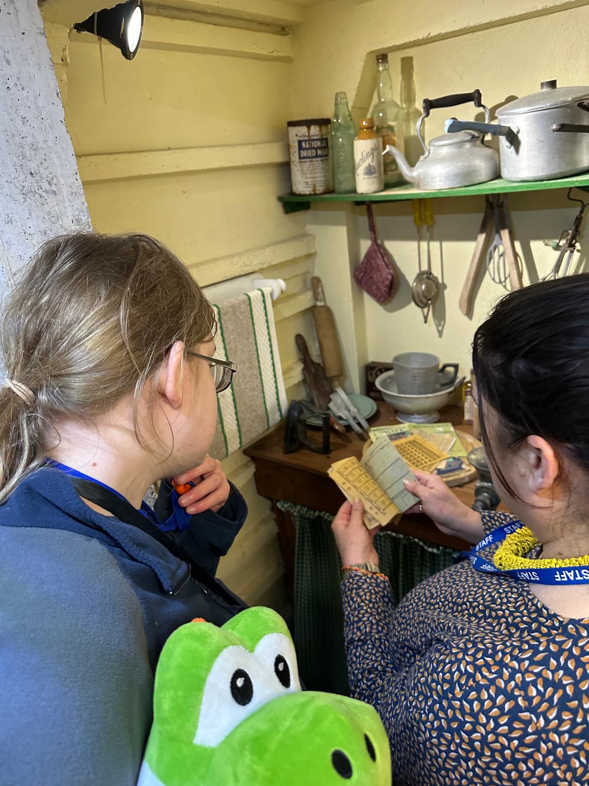A staff member shows a young person a small old pamphlet, in front of a display of domestic items from World War 2, including a kettle, sauce pan, bottles, bowls and other kitchen items.