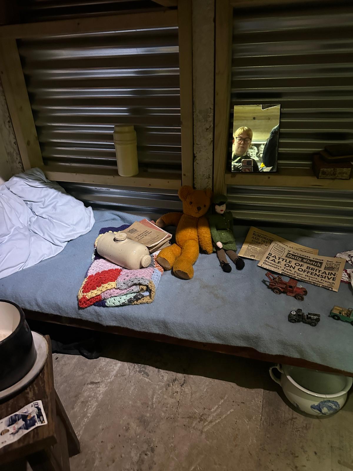 A simple child's bed with a blanket, some toys, newpapers, and a chamber pot under the bed.