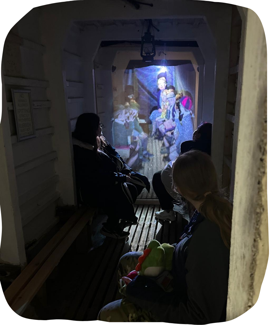 Young people sat on benches watching a projection of children in an air raid shelter.
