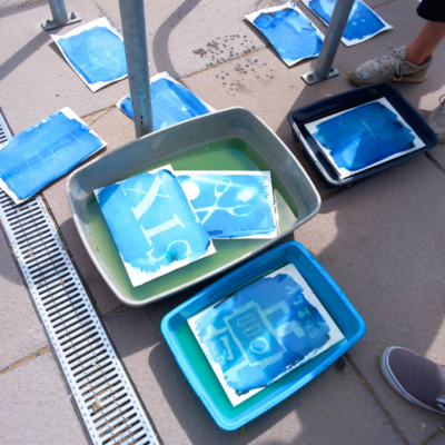 2 Sheets of paper undergoing the cyanotype process. They are blue where the liquid was applied, with white silhouettes of the shapes that were placed on top. They are in tubs of liquid outside, and some are drying on the floor.