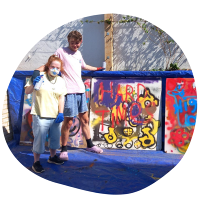 Two young people pose next to one of several large canvases with brightly coloured layered graffiti style spray art.