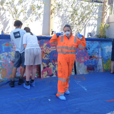 12 Young person wearing a protective jumpsuit, gloves and mask, doing two peace signs in front of several large canvases of spray art.