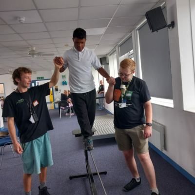 18 Young person walking on a tightrope a couple of feet above the ground. Two youth workers hold their hands to help them balance.